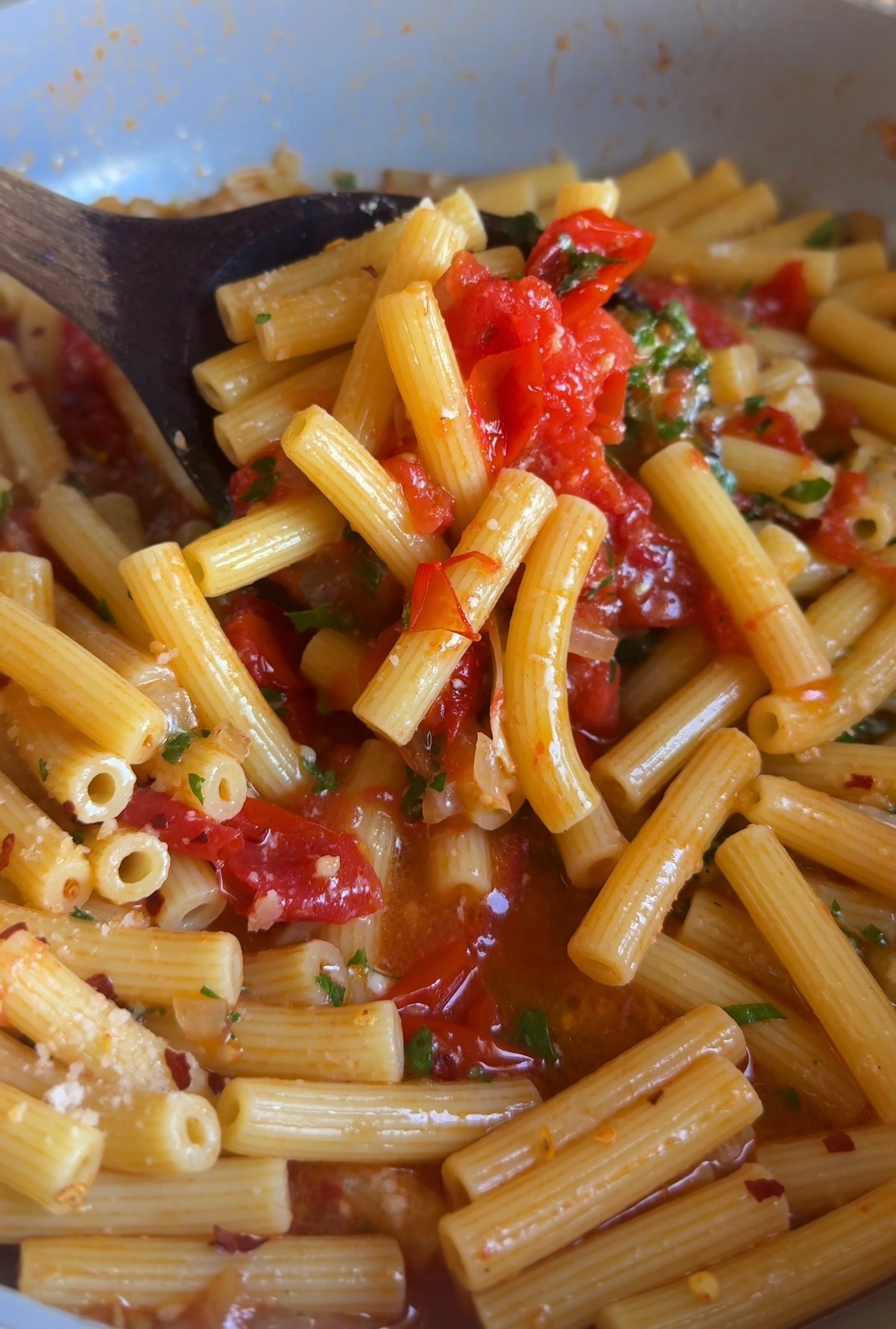 Rigatoni tossed with burst cherry tomatoes and herbs in a pan with a wooden spoon
