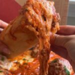 Hand lifting a saucy stuffed meatball cone with melted cheese pulling away