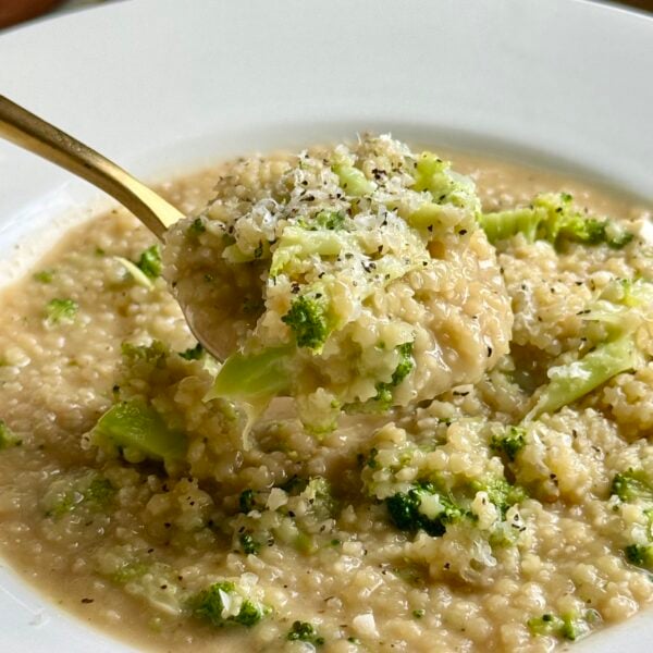 Gold spoon lifting creamy parmesan pastina studded with tender broccoli from a white bowl