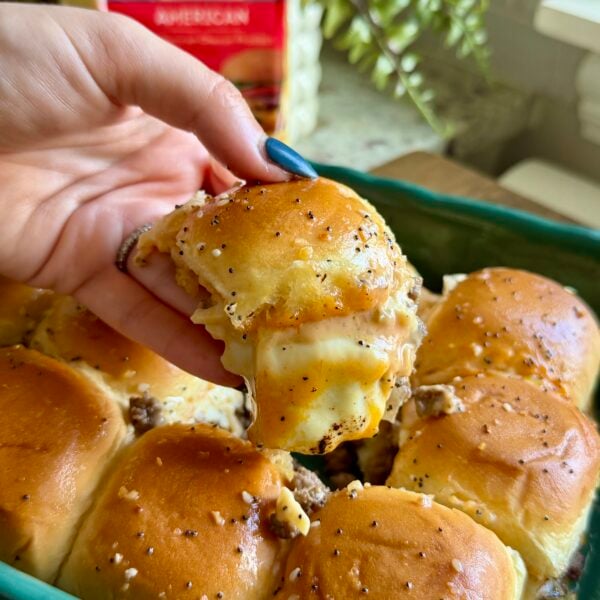 Hand lifting a cheeseburger slider with stretching cheese from a tray of poppyseed buns
