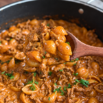 Wooden spoon lifting cheesy beef shells from creamy tomato sauce, flecked with parsley