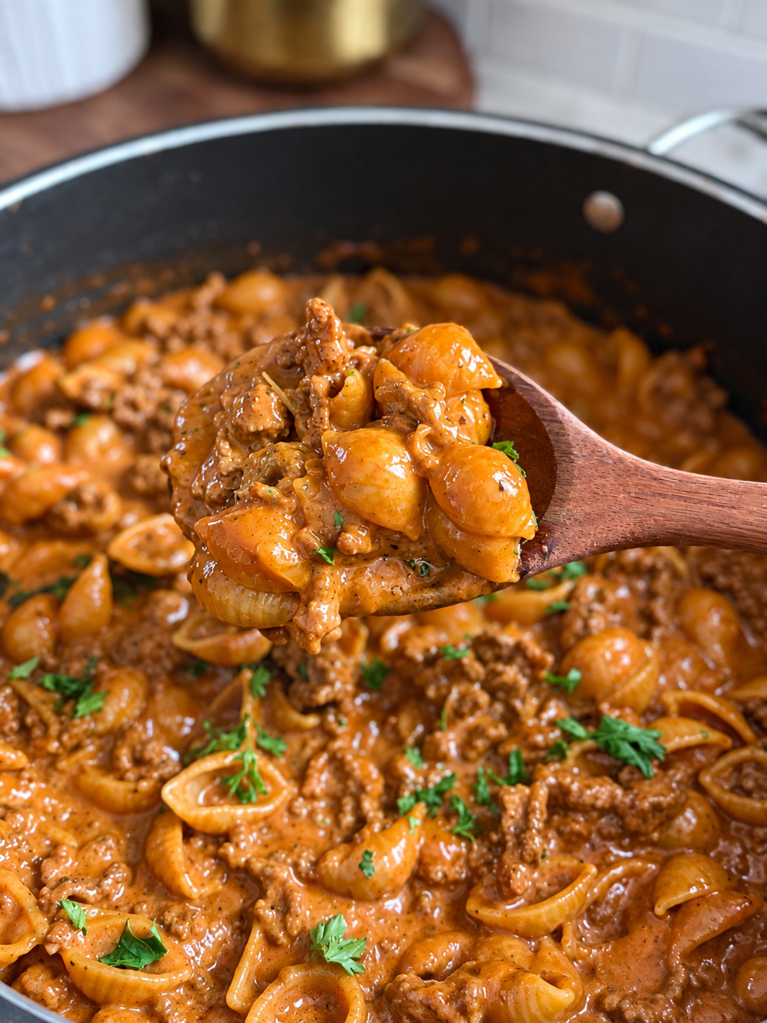 Wooden spoon lifting cheesy beef shells from creamy tomato sauce, flecked with parsley