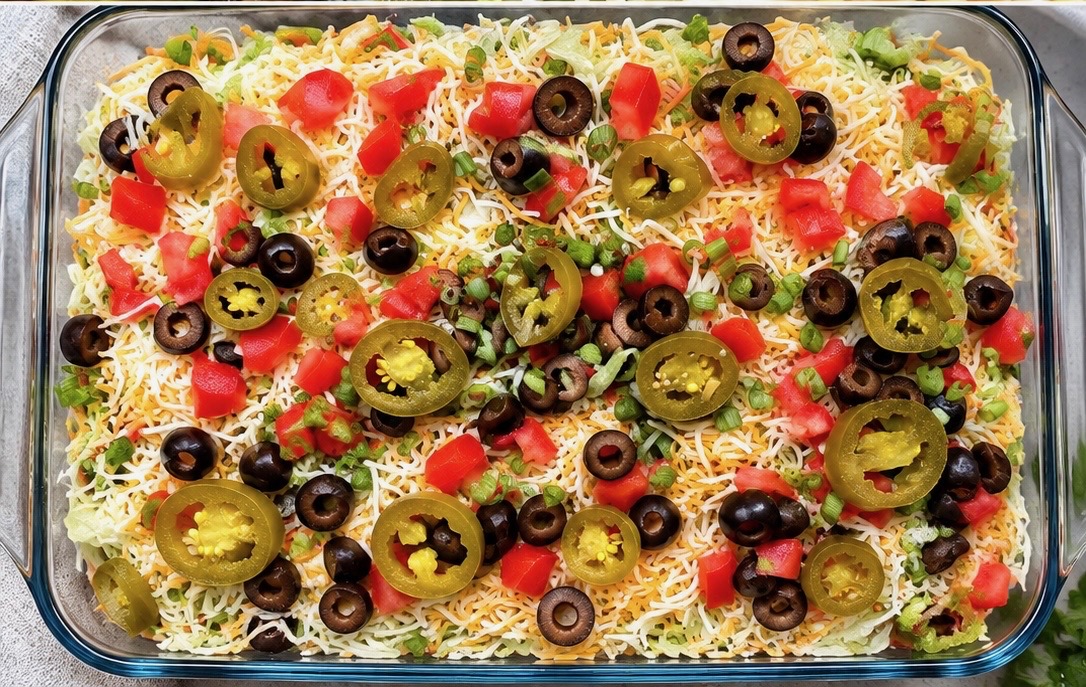 Glass dish of layered taco dip topped with cheese, tomatoes, olives and jalapenos