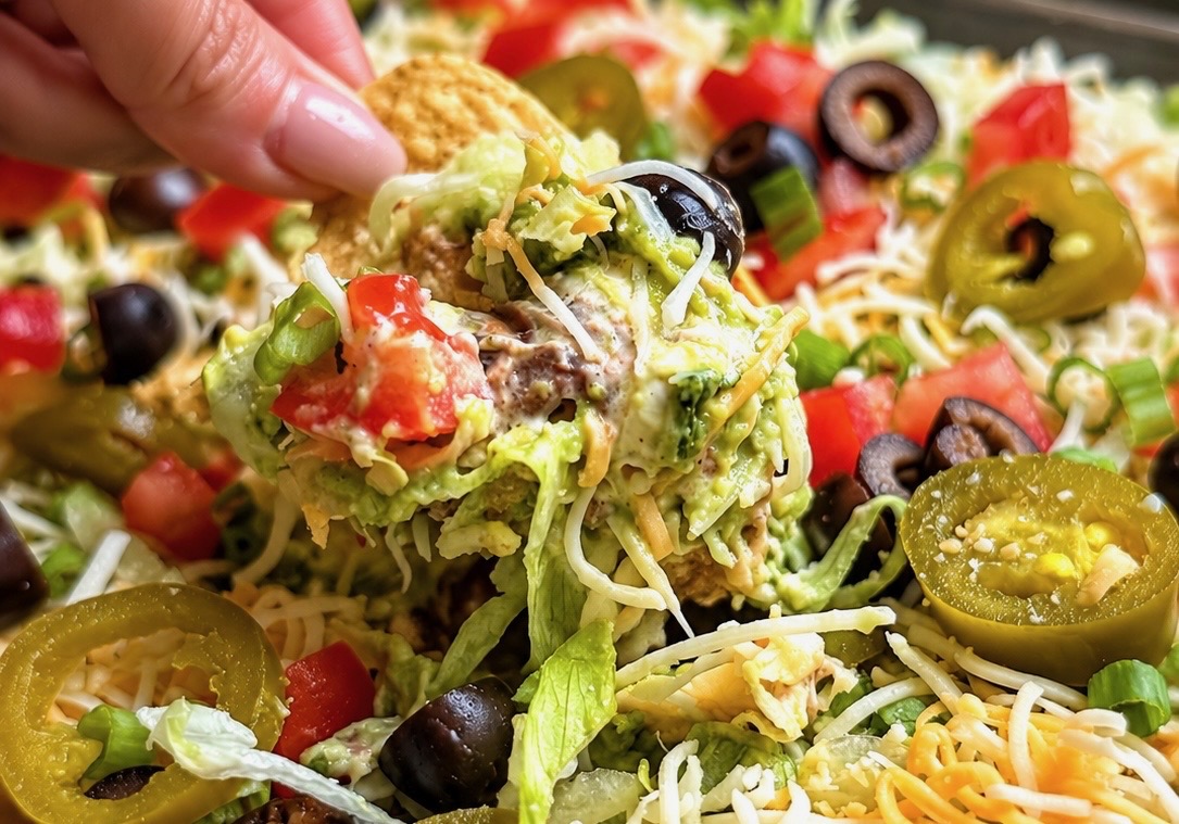 Hand lifting a loaded tortilla chip from layered taco dip with olives and jalapenos