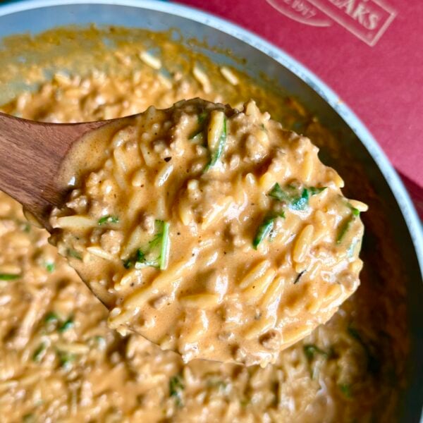 Wooden spoon lifting cheesy beef orzo flecked with spinach from the skillet