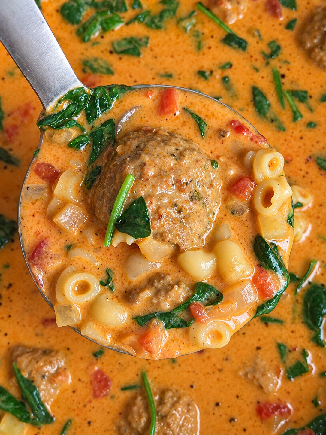 Ladle lifting a meatball and shells from creamy tomato soup with spinach