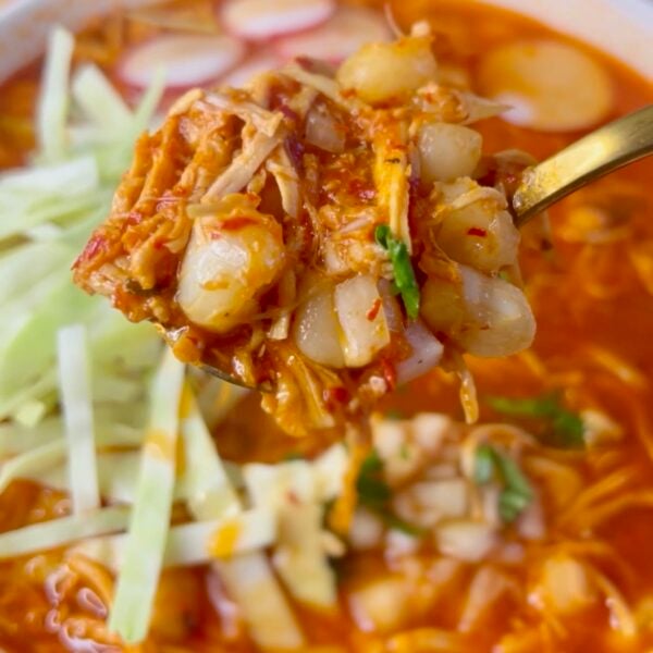 Spoon lifting shredded chicken and hominy from red pozole with cabbage and radish