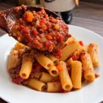 Wooden spoon ladling chunky bolognese sauce over rigatoni on a white plate