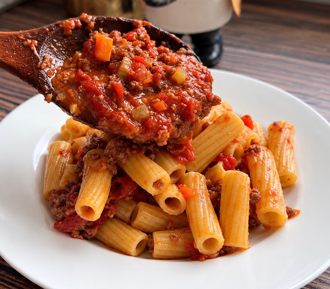 Wooden spoon ladling chunky bolognese sauce over rigatoni on a white plate