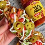 Hand holding taco stuffed potato skins topped with jalapeños, tomatoes and a sour cream drizzle