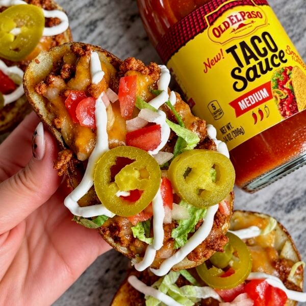Hand holding taco stuffed potato skins topped with jalapeños, tomatoes and a sour cream drizzle