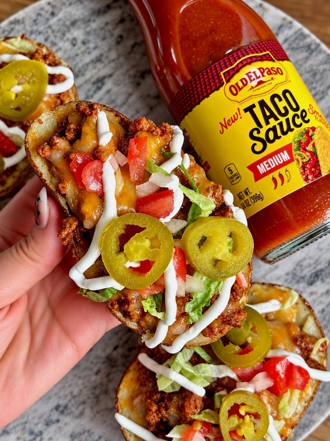 Hand holding taco stuffed potato skins topped with jalapeños, tomatoes and a sour cream drizzle