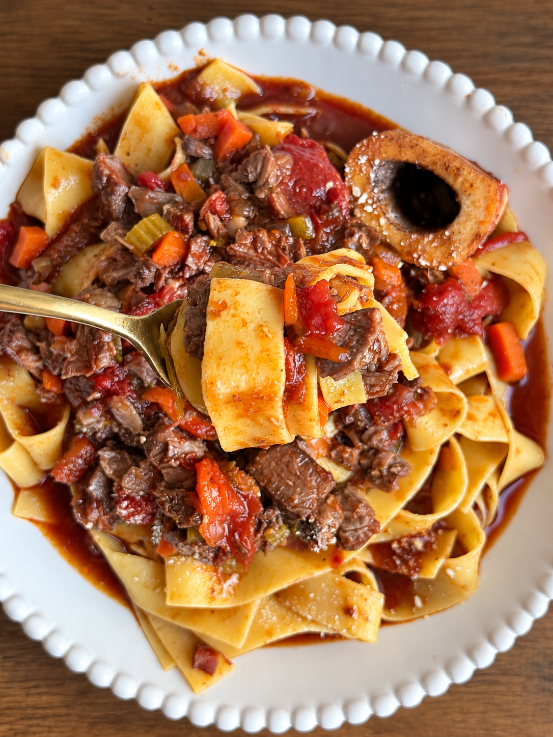 Gold fork lifting pasta with braised beef shank ragu beside a marrow bone